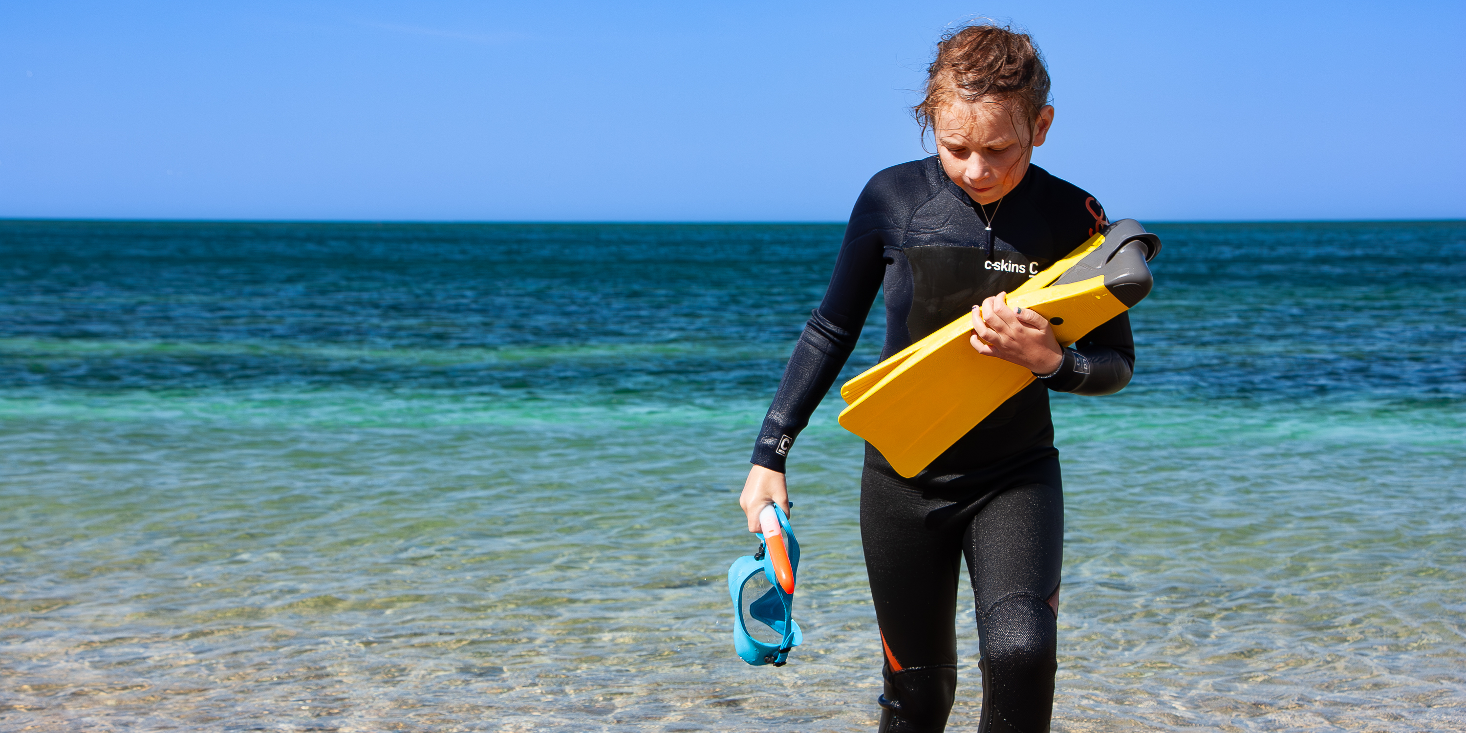 Photo of child walking up a beach holding snorkelling gear with blue-green sea behind